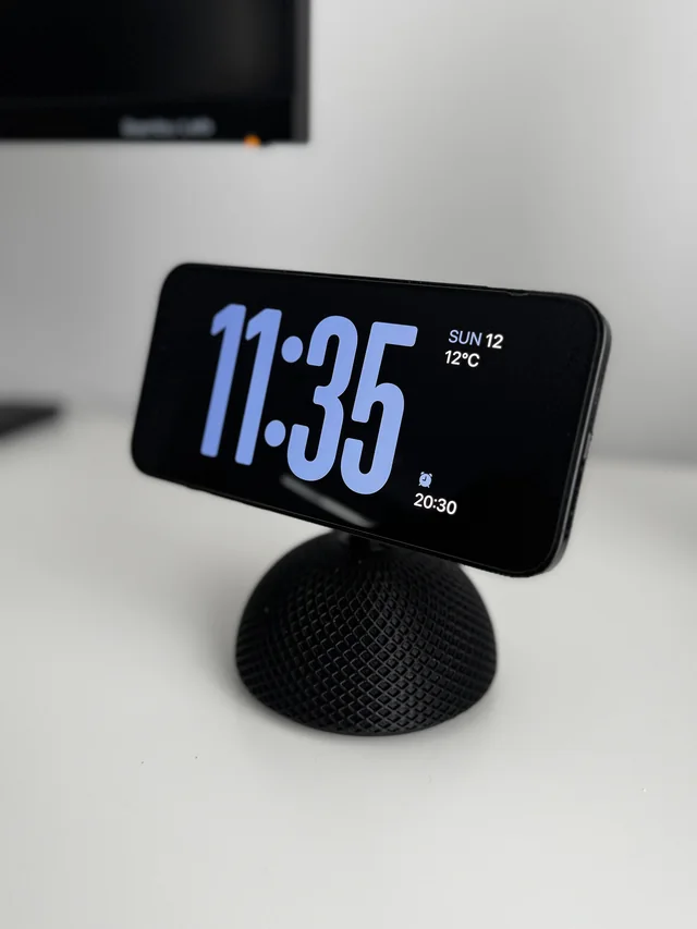 A MagSafe charger in a textured black 3D-printed housing, holding a phone horizontally on a white desk